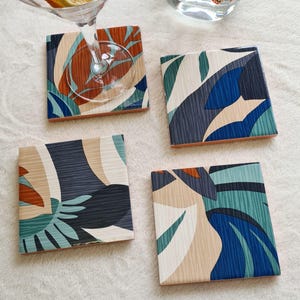 May include: Four square coasters featuring a graphic pattern of overlapping leaves in shades of navy blue, teal, light blue, and beige. The coasters have a textured surface finish. One coaster is partially covered by the base of a clear glass.