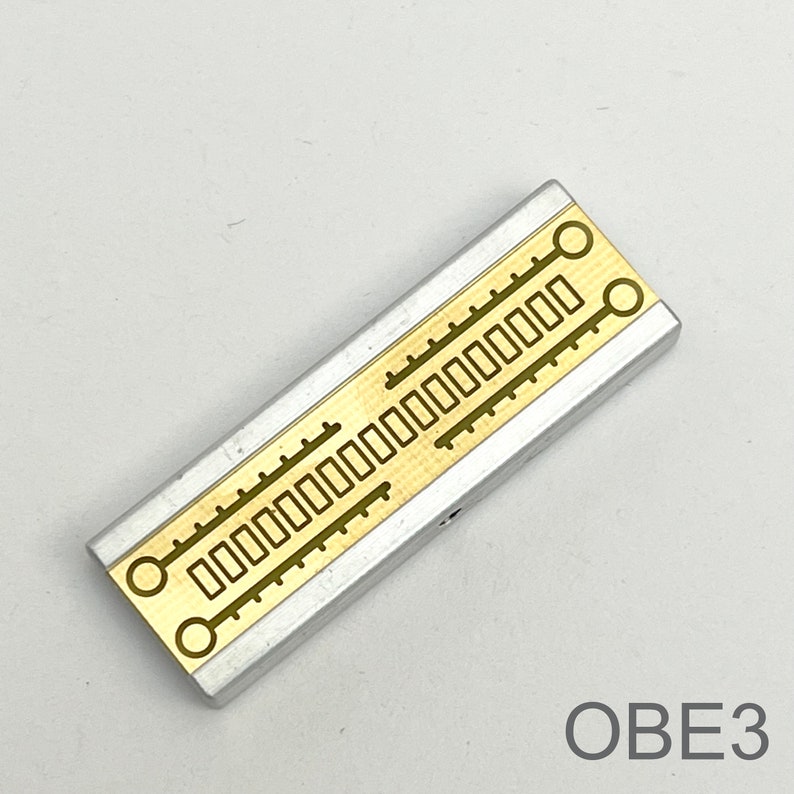 Peut inclure: Un composant &eacute;lectronique argent&eacute; et dor&eacute; avec une s&eacute;rie de d&eacute;coupes rectangulaires et circulaires. Le composant est &eacute;tiquet&eacute; "OBE3".