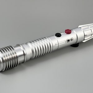 Puede incluir: Una r&eacute;plica de sable l&aacute;ser plateado con un agarre texturizado, botones negros y rojos y un soporte de hoja plateado.