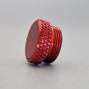 May include: A red metal cap with a textured, diamond-patterned grip and threaded sides. The cap has a flat, circular top and is designed to screw onto a corresponding fitting. The color is a vibrant, solid red.