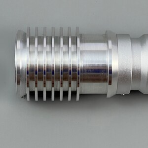 Puede incluir: Una extensi&oacute;n de sable de luz de metal plateado con una serie de anillos o ranuras a lo largo de la extensi&oacute;n.