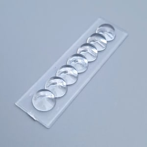 Könnte beinhalten: Nahaufnahme von sieben transparenten, gewölbten Klebepuffern, die in einer Reihe auf einer rechteckigen, transparenten Unterlage angeordnet sind. Die Puffer sind gleichmäßig verteilt und scheinen aus einem klaren, gelartigen Material zu bestehen. Der Hintergrund ist einfarbig hellgrau.