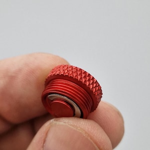 May include: A close-up of a red metal cap with a textured, ridged top and threaded sides. The cap has a small, circular, silver-colored component in the center. The cap is held between fingers against a blurred background.