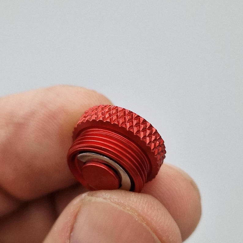 May include: A close-up of a red metal cap with a textured, ridged top and threaded sides. The cap has a small, circular, silver-colored component in the center. The cap is held between fingers against a blurred background.