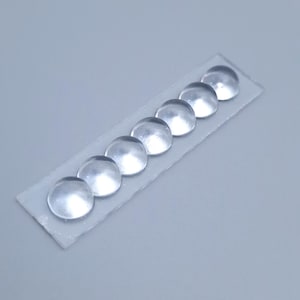 Könnte beinhalten: Sieben klare, gewölbte Glascabochons, jeweils etwa 1,3 cm im Durchmesser, sind auf einem dünnen, rechteckigen, durchscheinenden Klebestreifen angeordnet. Die Cabochons haben eine glatte, abgerundete Oberfläche vor einem hellgrauen Hintergrund.