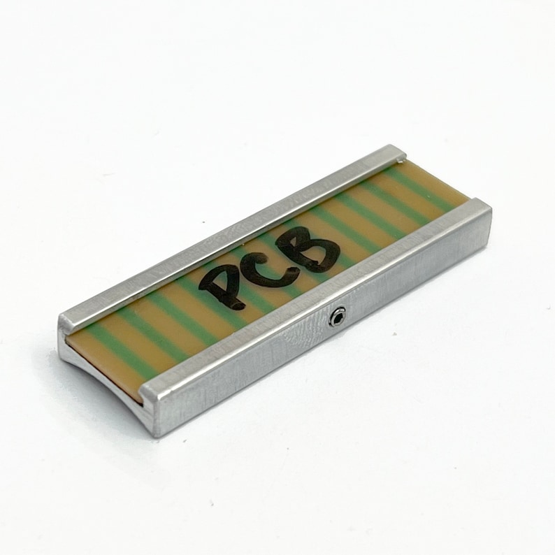 Peut inclure: Un cadre en m&eacute;tal argent&eacute; avec un circuit imprim&eacute; &agrave; rayures vertes et beiges &agrave; l'int&eacute;rieur. Le circuit imprim&eacute; porte l'inscription "PCB" &eacute;crite en noir.