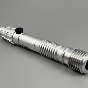 Puede incluir: Un accesorio de sable de luz plateado con una perilla negra y un dise&ntilde;o acanalado. El sable de luz est&aacute; hecho de metal y tiene una tapa de extremo negra.