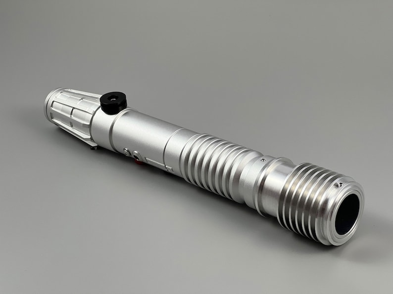 Puede incluir: Un accesorio de sable de luz plateado con una perilla negra y un dise&ntilde;o acanalado. El sable de luz est&aacute; hecho de metal y tiene una tapa de extremo negra.