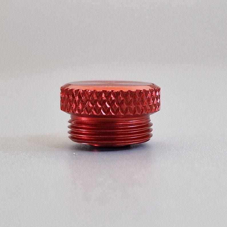 May include: A close-up of a red metal cap with a textured, diamond-patterned top and threaded sides. The cap is likely for a container or device, and it is set against a neutral background.
