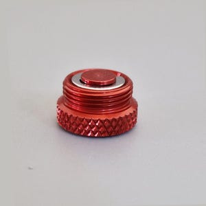 May include: A red metal cap with a textured, diamond-patterned base and a threaded body. The cap has a smaller, circular top with a silver ring around it. The object is likely a component or accessory.