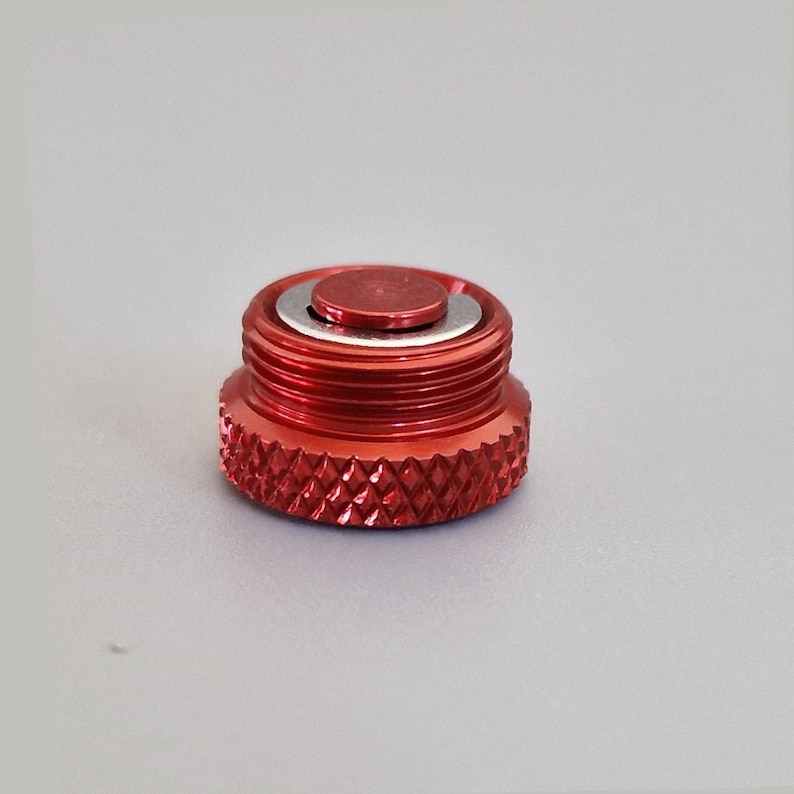 May include: A red metal cap with a textured, diamond-patterned base and a threaded body. The cap has a smaller, circular top with a silver ring around it. The object is likely a component or accessory.