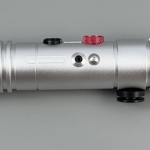 Puede incluir: Una r&eacute;plica de sable de luz plateado con detalles en negro y rojo. El sable de luz tiene una perilla negra en el extremo y una perilla roja en el lateral.