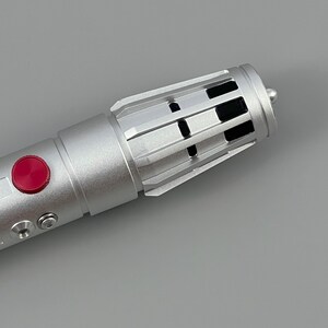 Puede incluir: Un sable de luz plateado con un bot&oacute;n rojo y respiraderos negros. El sable de luz est&aacute; hecho de metal y tiene un dise&ntilde;o elegante.