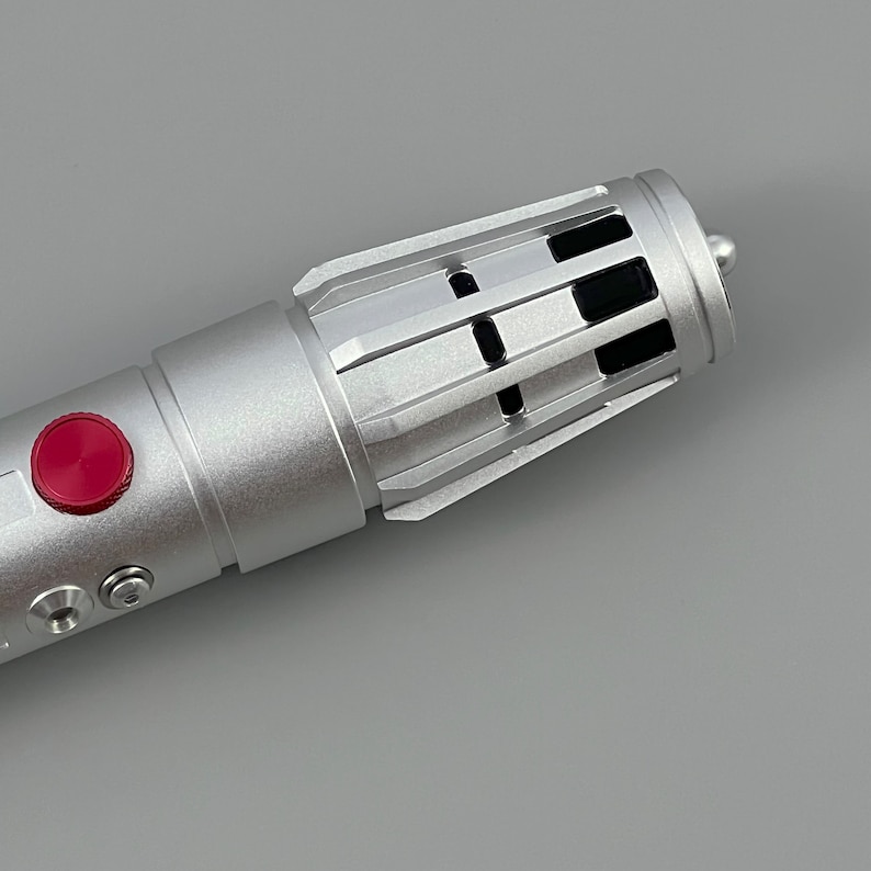 Puede incluir: Un sable de luz plateado con un bot&oacute;n rojo y respiraderos negros. El sable de luz est&aacute; hecho de metal y tiene un dise&ntilde;o elegante.