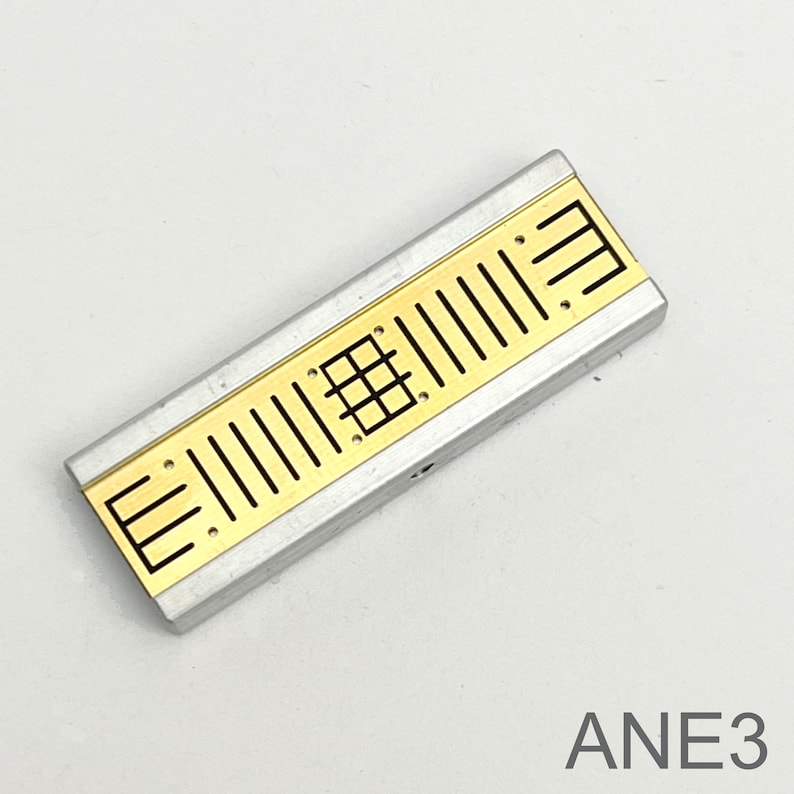Peut inclure: Un composant &eacute;lectronique rectangulaire argent&eacute; et dor&eacute; avec un motif en grille et le texte "ANE3" imprim&eacute; sur la surface blanche en dessous.