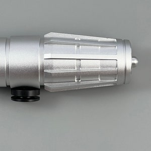 Puede incluir: Una empu&ntilde;adura de sable de luz de metal plateado con un dise&ntilde;o texturizado y estriado. La empu&ntilde;adura tiene una empu&ntilde;adura de goma negra y una punta plateada.