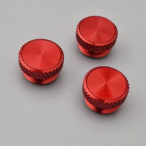 Könnte beinhalten: Drei rote, kreisförmige Metallobjekte mit einem strukturierten, geriffelten Rand und einer flachen, leicht konkaven Oberseite. Die Objekte sind gleichmäßig auf einem grauen Hintergrund angeordnet. Die Oberseiten haben ein radiales Muster, das von der Mitte ausgeht.