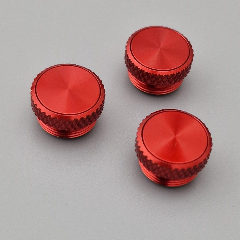 May include: Three red, circular metal objects with a textured, ridged edge and a flat, slightly concave top. The objects are evenly spaced against a gray background. The tops have a radial pattern emanating from the center.
