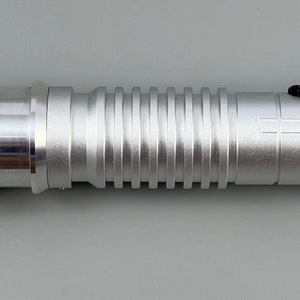 Puede incluir: Un sable de luz plateado con una superficie texturizada y estriada. El sable de luz est&aacute; hecho de metal y tiene un acento negro en el extremo.