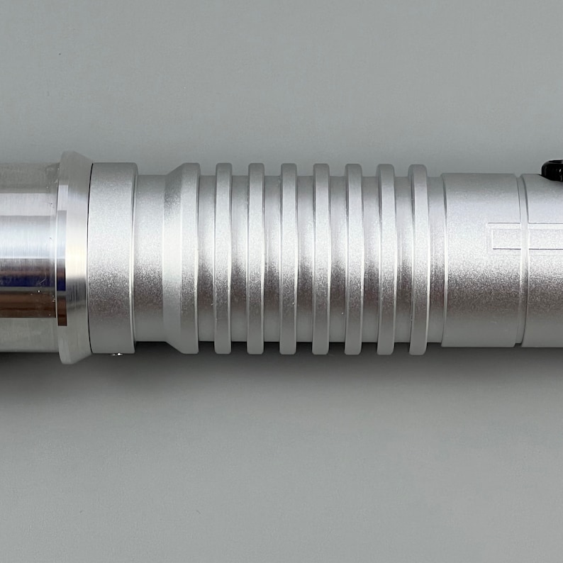 Puede incluir: Un sable de luz plateado con una superficie texturizada y estriada. El sable de luz est&aacute; hecho de metal y tiene un acento negro en el extremo.