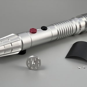 Puede incluir: Un sable de luz plateado con un bot&oacute;n negro y rojo, un emisor negro y plateado y una empu&ntilde;adura negra y plateada. El sable de luz est&aacute; desmontado y dispuesto sobre una superficie gris. Las piezas incluyen un emisor negro y plateado, una empu&ntilde;adura negra y plateada, una hoja negra y plateada y una celda de energ&iacute;a negra y plateada.