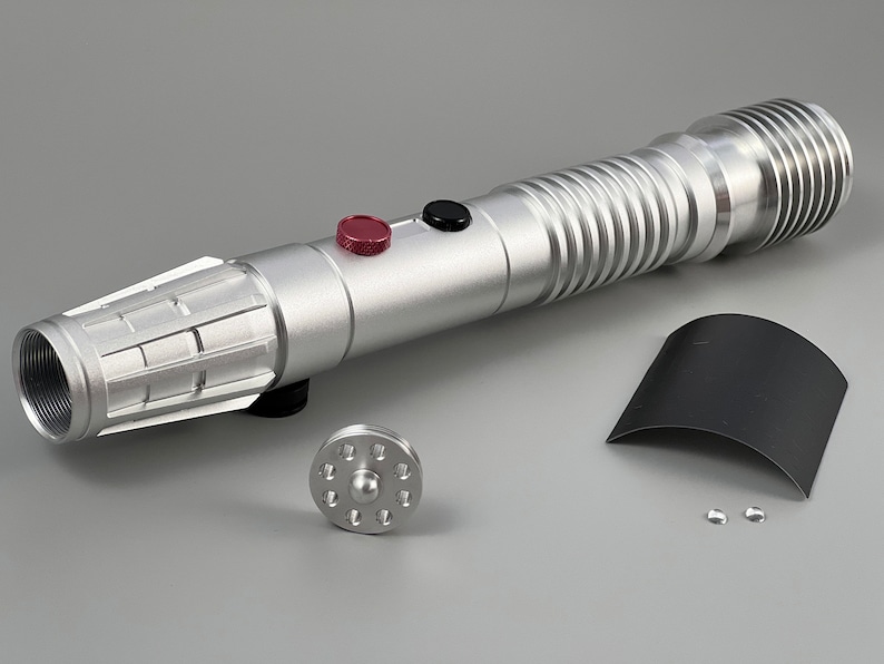 Puede incluir: Un sable de luz plateado con un bot&oacute;n negro y rojo, un emisor negro y plateado y una empu&ntilde;adura negra y plateada. El sable de luz est&aacute; desmontado y dispuesto sobre una superficie gris. Las piezas incluyen un emisor negro y plateado, una empu&ntilde;adura negra y plateada, una hoja negra y plateada y una celda de energ&iacute;a negra y plateada.