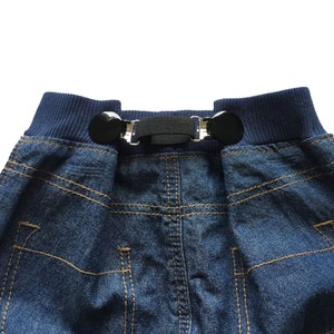 Könnte beinhalten: Eine blaue Jeans mit einem schwarzen elastischen Bund und zwei silbernen Verschlüssen. Der Bund hat einen dunkelblauen Saum.