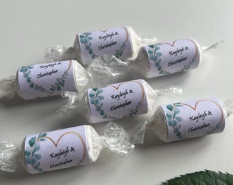 May include: Six wrapped candies with white labels featuring the names "Kayleigh & Christopher" and a gold heart design with green floral accents. The candies are on a white surface with a green leaf.