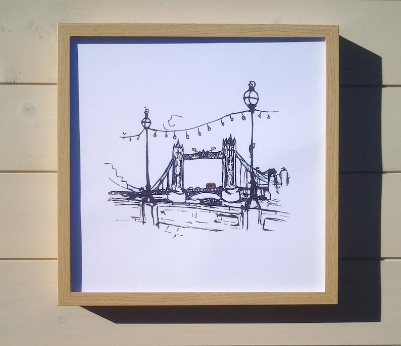 May include: A black and white line drawing of Tower Bridge in London, England. The bridge is depicted with string lights strung across the top. The drawing is framed in a light wood frame.