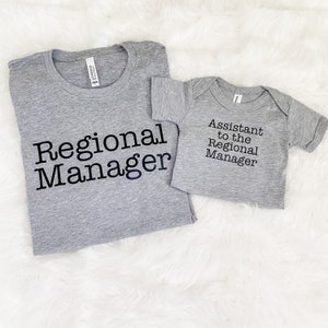 May include: Two gray t-shirts, one adult and one infant, with black text. The adult shirt reads "Regional Manager," and the infant shirt says "Assistant to the Regional Manager." Both shirts are on a white, fluffy surface.