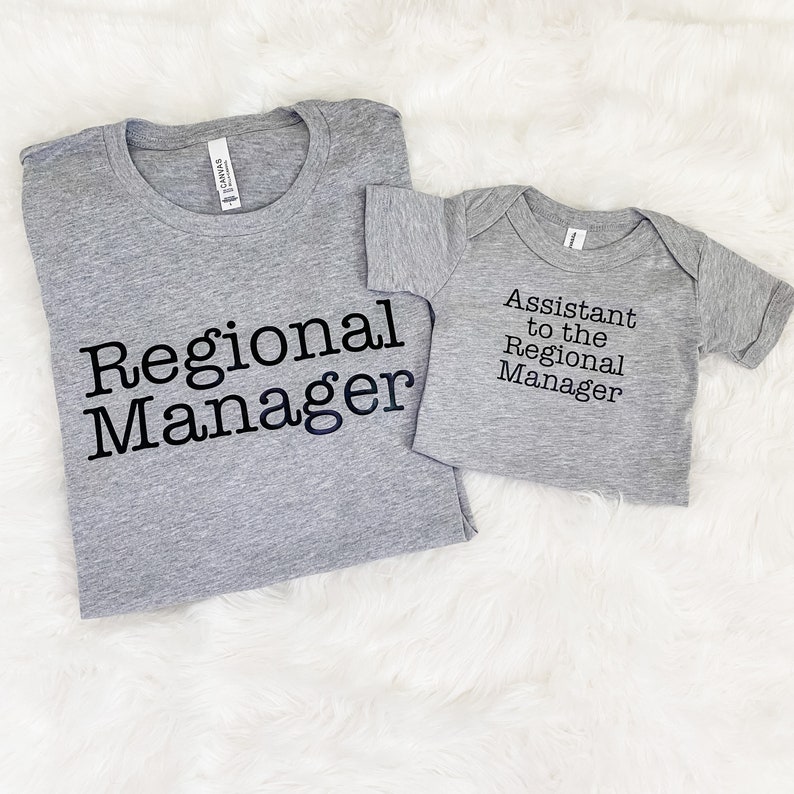 May include: Two gray t-shirts, one adult and one infant, with black text. The adult shirt reads "Regional Manager," and the infant shirt says "Assistant to the Regional Manager." Both shirts are on a white, fluffy surface.