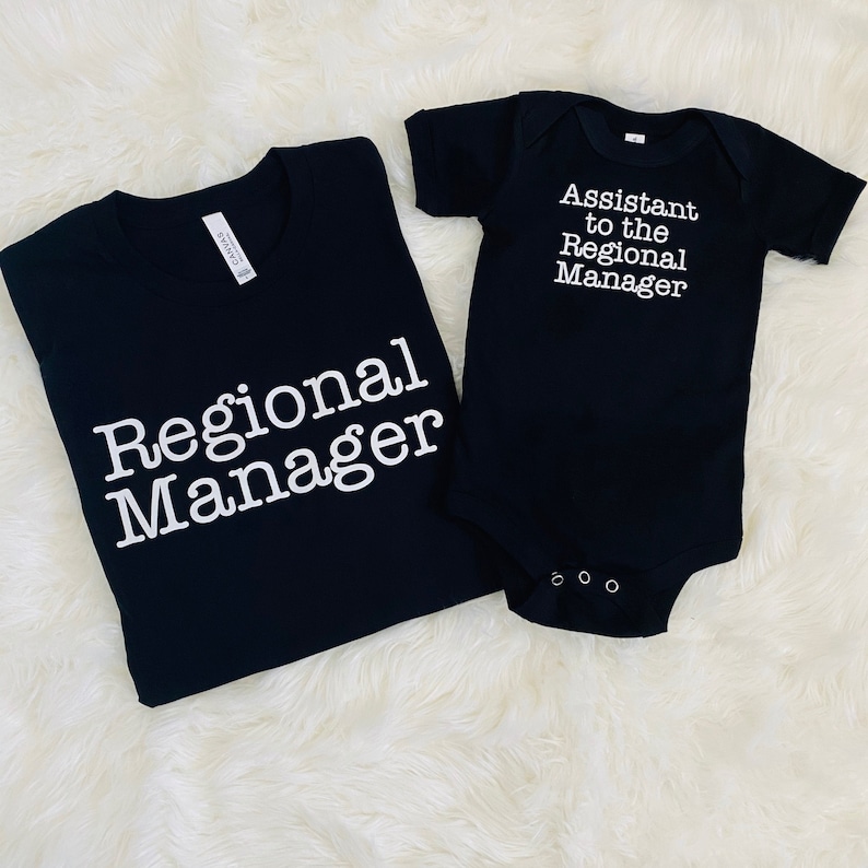 May include: Black t-shirt with the text "Regional Manager" and a black baby onesie with the text "Assistant to the Regional Manager" in white. The items are displayed on a white, fluffy surface.