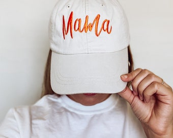 May include: A white baseball cap with the word "Mama" embroidered in orange and red.