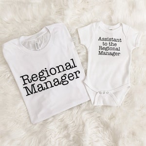 May include: White t-shirt with the text "Regional Manager" and a white baby onesie with the text "Assistant to the Regional Manager" in black. The clothing items are displayed on a white, fluffy surface.