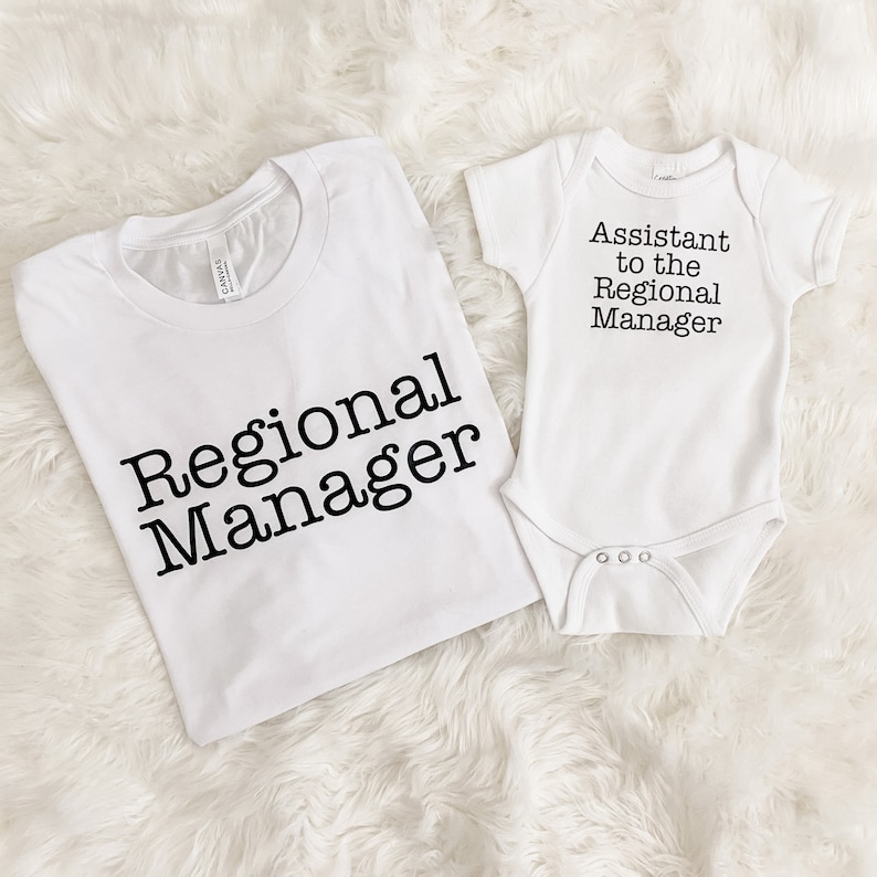May include: White t-shirt with the text "Regional Manager" and a white baby onesie with the text "Assistant to the Regional Manager" in black. The clothing items are displayed on a white, fluffy surface.