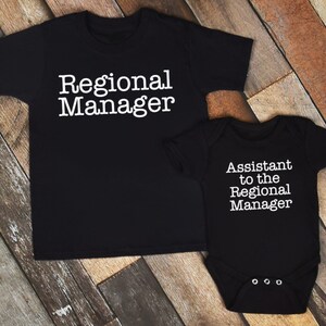 May include: Black t-shirt and onesie set. The t-shirt reads "Regional Manager" in white text. The onesie reads "Assistant to the Regional Manager" in white text. The set is displayed on a wooden surface.