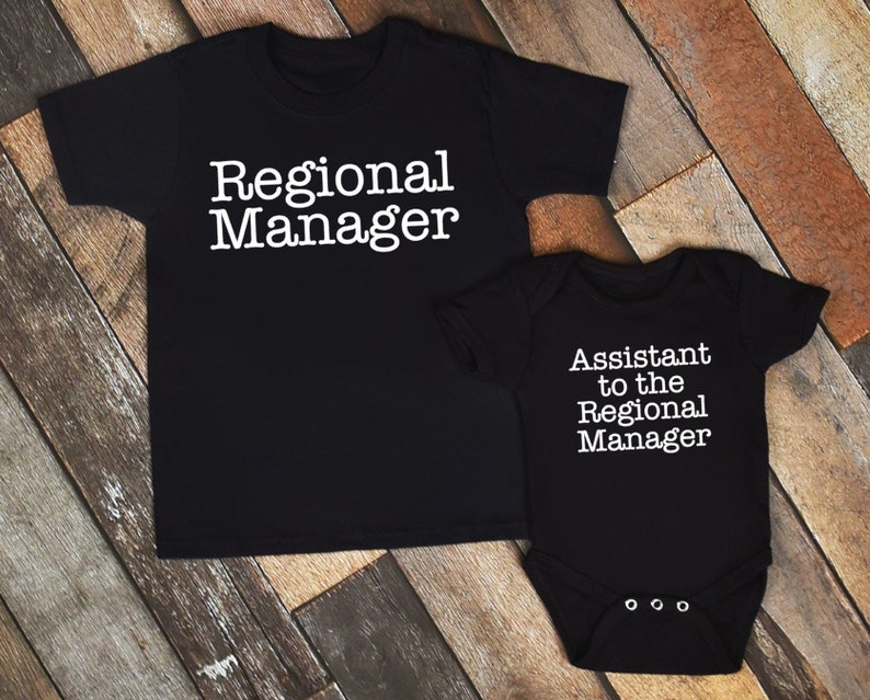 May include: Black t-shirt and onesie set. The t-shirt reads "Regional Manager" in white text. The onesie reads "Assistant to the Regional Manager" in white text. The set is displayed on a wooden surface.