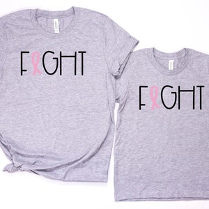 May include: Two grey t-shirts with the word "FIGHT" printed in black with a pink ribbon in the middle of the word. The shirts are a matching set.