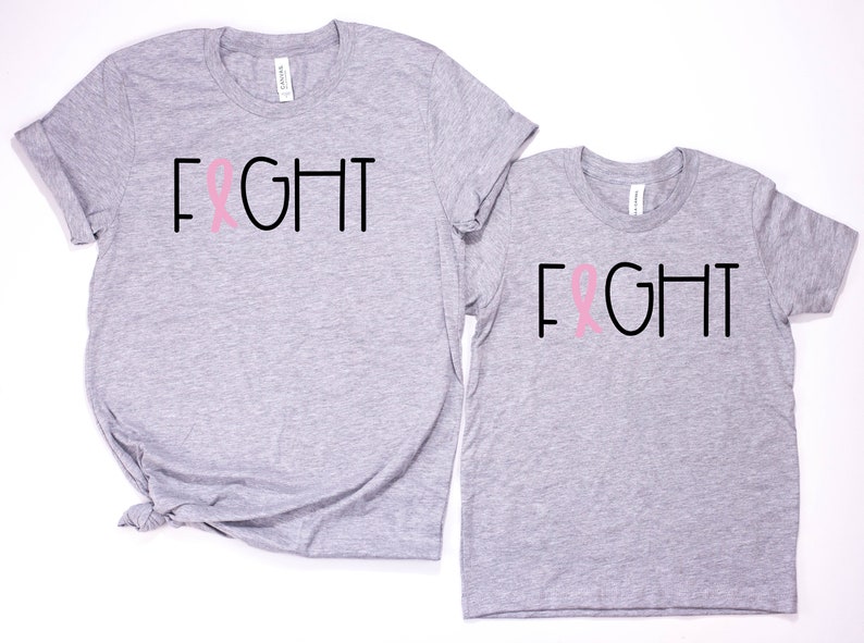 May include: Two grey t-shirts with the word "FIGHT" printed in black with a pink ribbon in the middle of the word. The shirts are a matching set.