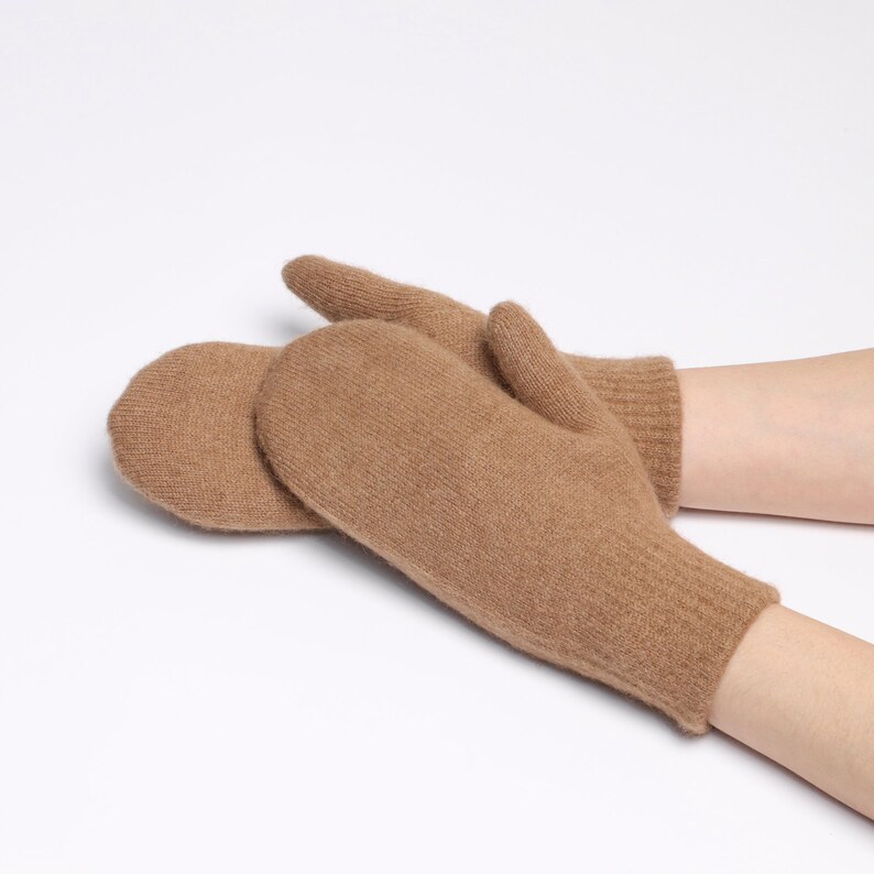 May include: A pair of tan-colored knit mittens featuring a soft, textured surface and ribbed cuffs. The mittens are designed with a rounded shape for the fingers and a separate section for the thumb, providing a simple and functional hand covering.
