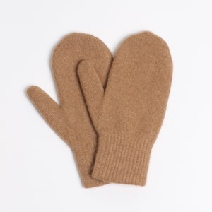 May include: A pair of tan-colored knit mittens featuring a soft, textured surface and a ribbed cuff design. The mittens are positioned overlapping each other, showcasing the consistent light brown yarn construction and the rounded shape of the fingers.