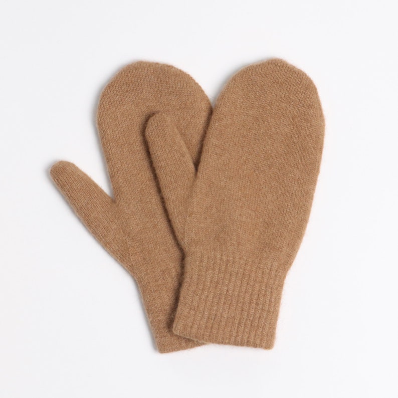May include: A pair of tan-colored knit mittens featuring a soft, textured surface and a ribbed cuff design. The mittens are positioned overlapping each other, showcasing the consistent light brown yarn construction and the rounded shape of the fingers.