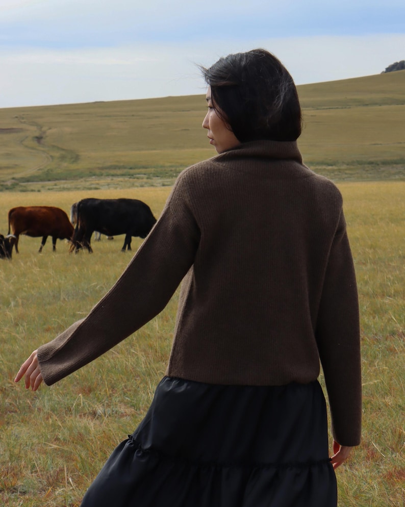 May include: A woman wearing a brown turtleneck sweater and a black tiered skirt stands in a field with cows in the background.