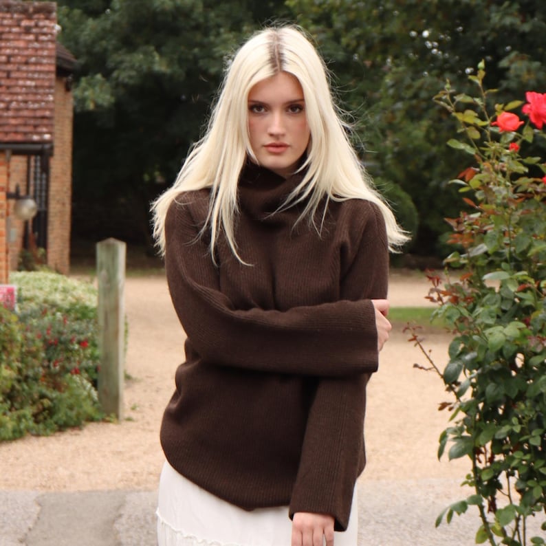 May include: A woman with long blonde hair is wearing a brown turtleneck sweater. The sweater has a relaxed fit and long sleeves. She is standing in front of a green bush.