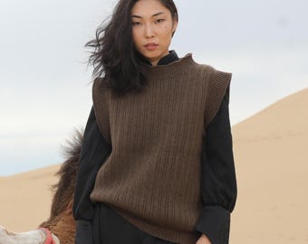 May include: A woman wearing a brown ribbed knit sleeveless sweater with a black mock neck shirt underneath. She is standing in a desert setting.
