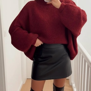Peut inclure: Un pull en tricot bordeaux surdimensionné avec un col montant et des manches longues. Associé à une mini-jupe en cuir noire et des bottes noires montantes. La tenue est présentée dans un couloir avec une rampe blanche.