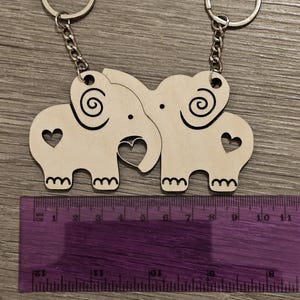 May include: Two wooden elephant keychains with a heart cutout and a swirl design. The keychains are attached to silver key rings.