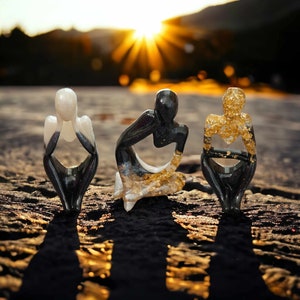 May include: Three small abstract sculptures made of black and white resin with gold accents. The sculptures are sitting on a wooden surface.
