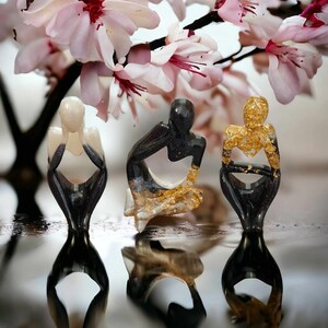 May include: Three small, abstract sculptures in a seated pose. The sculptures are made of black and white resin with gold accents. The sculptures are sitting on a reflective surface.