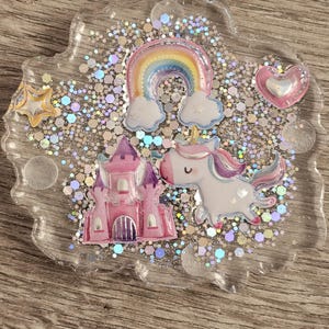 Pode incluir: Um descanso de copo de resina transparente com um castelo rosa, um unicórnio branco, um arco-íris e um coração rosa. O descanso de copo está cheio de glitter iridescente.